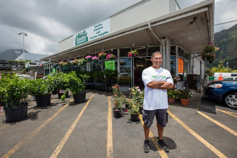 Smiling man in white t-shirt standing in front of Koolau Farmers store, with potted plants behind him