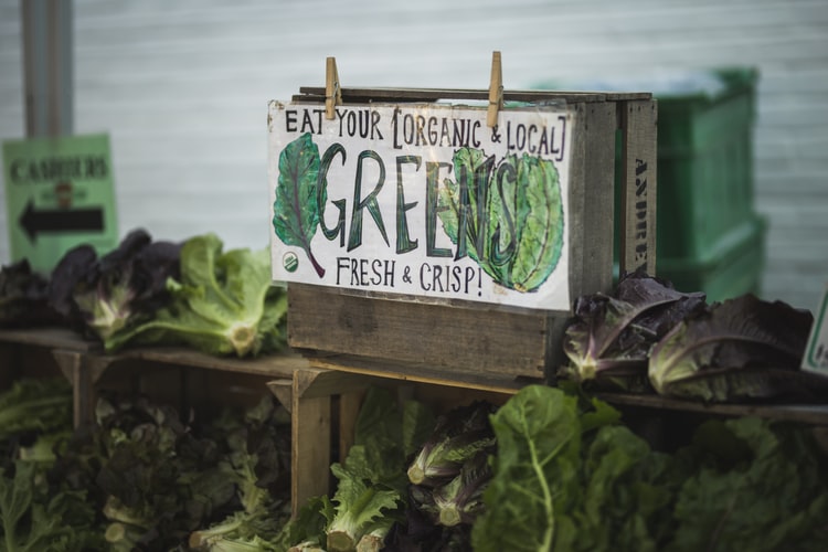 Wooden shelves filled with dark green vegetables and a box with "Eat Your Greens" sign on the front